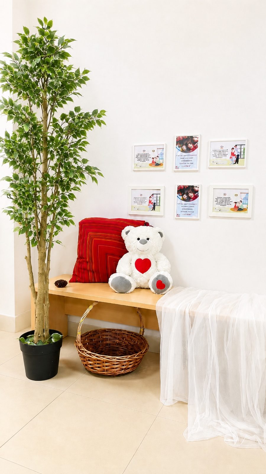 A quiet corner at Mosaic: framed prints above a light wood bench with a red cushion, a large teddy bear, a woven basket and a tall potted ficus