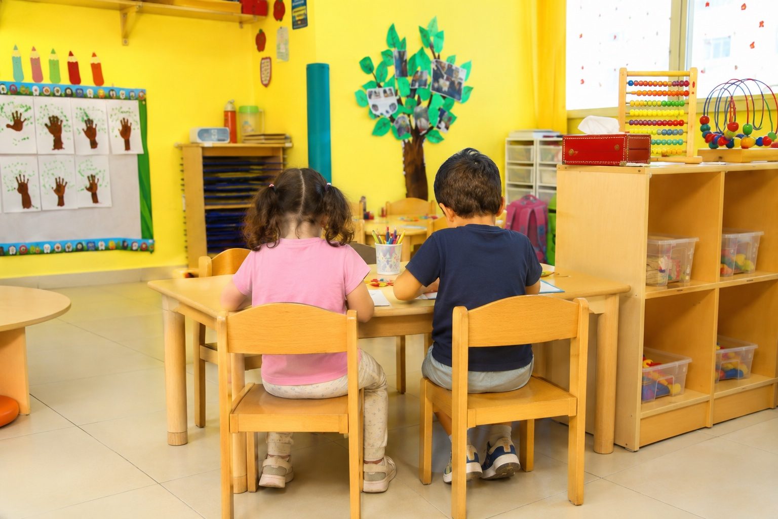 Two children working together at a low wooden table beside open shelving